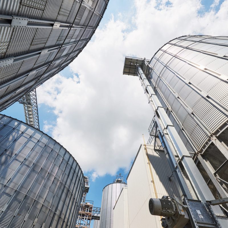 Agricultural Silos. Building Exterior. Storage and drying of grains, wheat, corn, soy, sunflower against the blue sky with white clouds Agricultural Silos. Building Exterior. Storage and drying of grains, wheat, corn, soy, sunflower against the blue sky with white clouds.